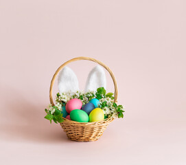Easter eggs with rabbit ears and flowers in a basket on pink background. Easter greeting card with space for text. Cute holiday decor.
