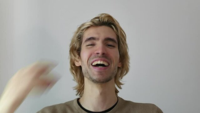 Adult young man with bleached blonde hair laughing in front of camera, wall in background, 4k resolution