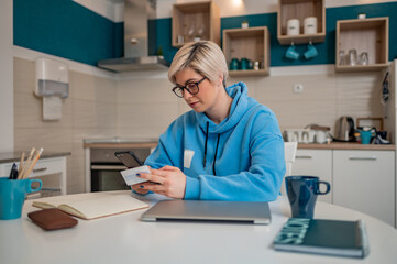 Obraz na płótnie Canvas Woman with a blond short hair holding credit card and using smartphone at home