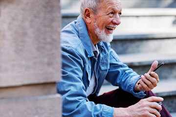 A senior man laughing at messages on the phone while sitting on stirs outside.