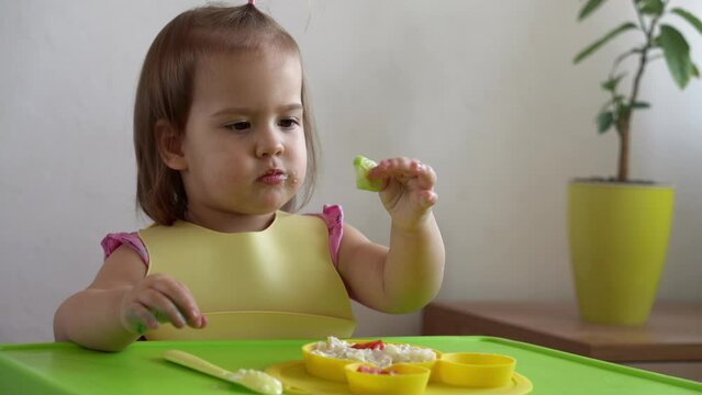 Close up of lovely baby girl eating have breakfast eggs vegetables sausage by herself at home. Adorable possitive happy toddler child eats with dirty face at table indoor. Food, nutrition healthy life