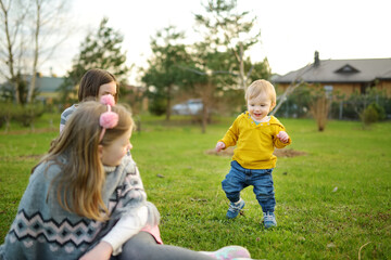 Fototapeta premium Two big sisters and their toddler brother having fun outdoors. Two young girls holding baby boy on summer day. Children with large age gap. Big age difference between siblings.