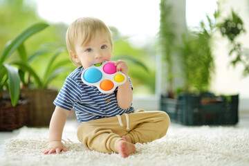 Toddler boy playing with rainbow pop-it fidget toy at home. Small child with stress and anxiety relief fidgeting game. - obrazy, fototapety, plakaty