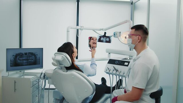 Young woman sitting on chair, looking at mirror, smiling after dentist procedure. Doctor and patient exploring teeth with mirror after treating in dental clinic. Concept of dentistry