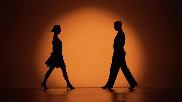 Couple of dancers approach each other and begin to dance Argentine tango. Elements of latin ballroom dance in studio with orange brown background. Dark silhouettes. Slow motion ready, 4K at 59.94fps.