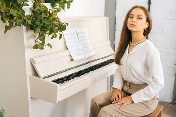 Portrait of successful young woman pianist looking at camera sitting at classical white piano in classroom. Satisfied attractive female musician performer preparing for classes at home studio.