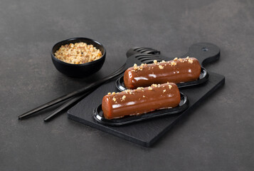 Chocolate walnut cake in the form of a cylindrical bar, with mirror glaze on a serving board on a dark gray background
