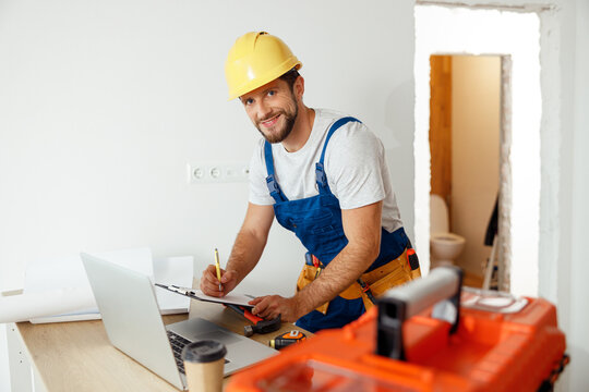 Smiling handyman worker in uniform and hard hat using laptop, writing on a clipboard, looking at camera while standing indoors