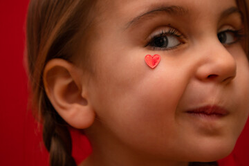 A funny little girl looks into the camera, with her heart on her cheek, with pigtails on a red background.