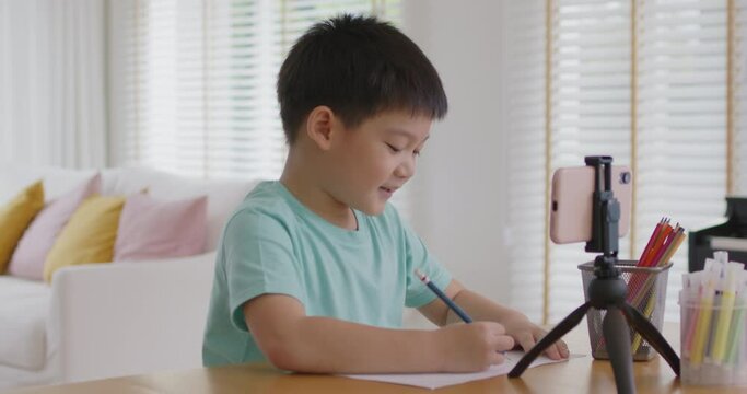 Asia cute smart boy alpha gen z kid enjoy fun hobby idea paint pencil color game on paper at online day care play school by vlog talk video call app. Young son study class tutor teach at home table.