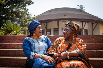 Two African women are conversing sitting on the stairs, they are both dressed in traditional clothes