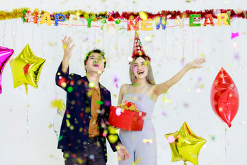 Millennial young lover couple Asian husband in casual suit and beautiful wife smiling holding red wrapped present gift box celebrating happy new year party in full decorated room with star balloons