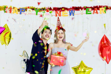 Millennial young lover couple Asian husband in casual suit and beautiful wife smiling holding red wrapped present gift box celebrating happy new year party in full decorated room with star balloons