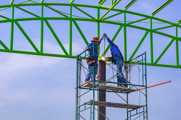 Two Asian welders on scaffolding are welding metal roof structure of industrial building against blue sky background 