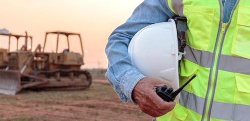 An engineer or worker wearing a safety vest holds a radio transmitter and a white helmet in his hands. for safety working on sunset background and crawler bulldozer