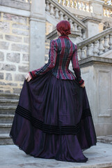 Full length portrait of red-haired woman wearing a historical victorian gown costume, walking around beautiful location with  Gothic stone architecture. - obrazy, fototapety, plakaty