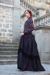 Fototapeta na wymiar Full length portrait of red-haired woman wearing a historical victorian gown costume, walking around beautiful location with Gothic stone architecture.