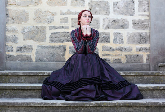 Full length portrait of red-haired woman wearing a historical victorian gown costume, walking around beautiful location with  Gothic stone architecture.