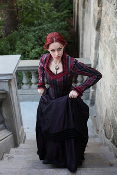 Full length portrait of red-haired woman wearing a historical victorian gown costume, walking around beautiful location with  Gothic stone architecture.