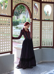 Full length portrait of red-haired woman wearing a historical victorian gown costume, walking around beautiful location with  Gothic stone architecture.