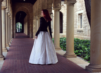 Full length portrait of red-haired woman wearing a  beautiful gothic gown costume, walking around  location with  romantic castle stone architecture background.