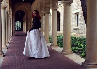 Full length portrait of red-haired woman wearing a  beautiful gothic gown costume, walking around  location with  romantic castle stone architecture background.