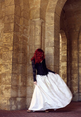 Full length portrait of red-haired woman wearing a  beautiful gothic gown costume, walking around  location with  romantic castle stone architecture background.