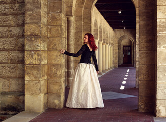 Full length portrait of red-haired woman wearing a  beautiful gothic gown costume, walking around  location with  romantic castle stone architecture background.