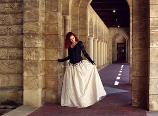 Naklejka premium Full length portrait of red-haired woman wearing a beautiful gothic gown costume, walking around location with romantic castle stone architecture background.