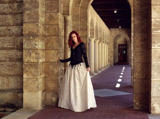 Naklejka premium Full length portrait of red-haired woman wearing a beautiful gothic gown costume, walking around location with romantic castle stone architecture background.