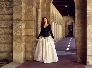 Full length portrait of red-haired woman wearing a  beautiful gothic gown costume, walking around  location with  romantic castle stone architecture background.