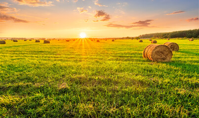 Scenic view at picturesque burning sunset in a green shiny field with hay stacks, bright cloudy sky , golden sun rays and road leading far away, summer valley landscape