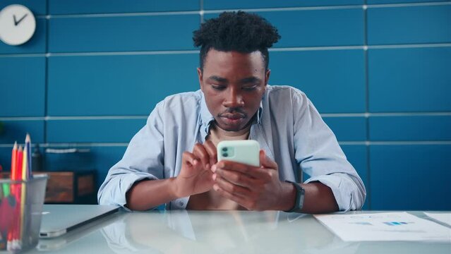 Young successful ethnic African American man published post on social networks via mobile phone doing blogging or internet journalism broadcasting online in print sits at desk in home office