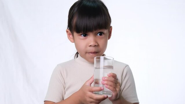 Cute little asian girl drinking water from a glass on white background in studio. Good healthy habit for children. Healthcare concept