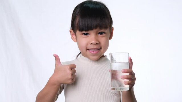 Cute little asian girl holding a glass of water and showing thumbs up on white background in studio. Good healthy habit for children. Healthcare concept