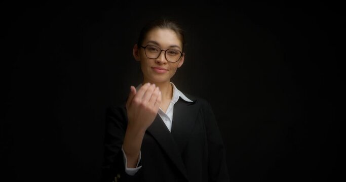 Businesswoman in glasses and a black jacket shows a come here hand gesture. A young Asian woman beckons with inviting gesture invites you to come . Isolated on a black background
