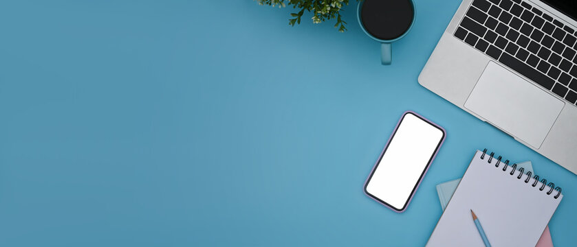 Smart phone, laptop computer, coffee cup and notebook on blue background with copy space.