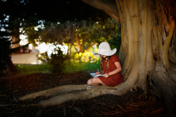 Fototapeta na wymiar A cute european kid girl in a dress and a big hat sits near a big tree in the park. A child among the big roots of a tree writes poetry, a girl makes notes