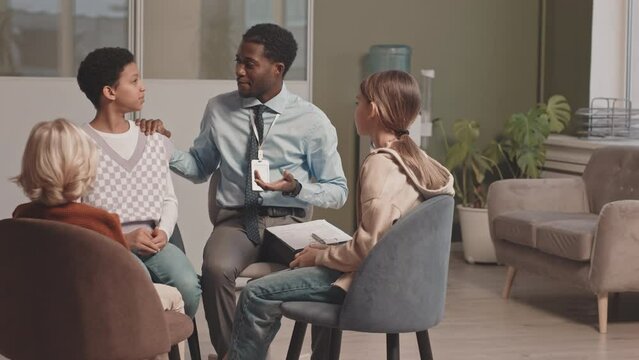 Medium slowmo shot of group of three diverse elementary kids and African-American male psychologist having conversation during therapy session, sitting in circle at modern doctors office