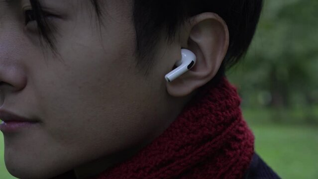 A man listens to music in wireless headphones. Young Asian man wearing headphones outdoors. Wireless earphone in the ear close-up