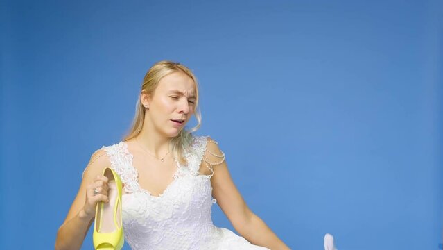 Tortured young beautiful blonde woman in a wedding white dress and veil on a blue background, takes off her shoes. The girl looks into the camera. Wedding