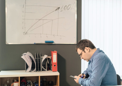 Man Employee In Blue Shirt And Glasses Sitting At The Desk In Office With Smartphone Using Digital Technology Applications For Business Development Against White Board With The Graph On Background. 