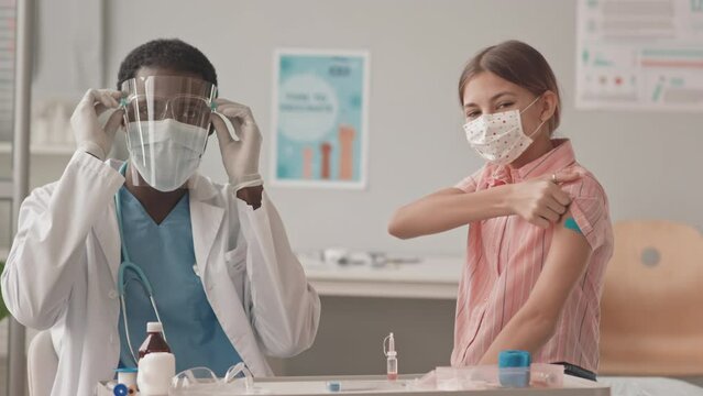 Medium slowmo portrait of masked African-American doctor and Caucasian schoolgirl looking at camera after Covid-19 vaccine injection. Girl showing band aid on her shoulder and smiling