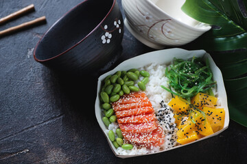 Poke with salmon, mango, edamame and seaweed in a carton delivery container, high angle view on a dark-brown stone surface, studio shot