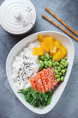 Poke bowl with rice, salmon fillet, edamame beans and mango, vertical shot on a grey concrete background, above view
