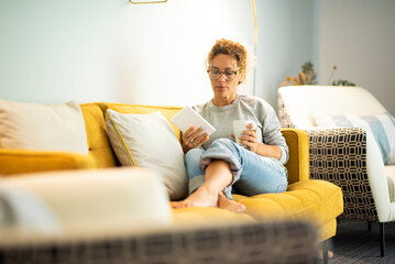 Confident caucasian adult woman sitting on the sofa reading a book using electronic device tablet reader. Pretty female people have relax and enjoy leisure activity at home laying on the couch