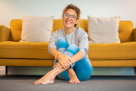 Adult Woman Smiling Sitting At Home Onthe Floor Against A Yelow Sofa. Happy Female People Have Relax Leisure Activity In Living Room. Young Lady Enjoyed Time Comfortably On The Carpet