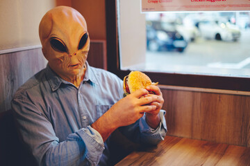 Alien with human clothes eating hamburger sandwich inside a fast food sitting at the table alone....