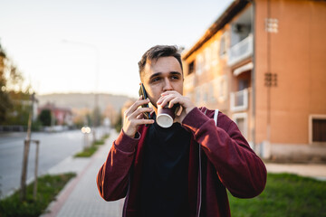 Front view of one adult young caucasian man walking in front of street in town in autumn evening holding and using mobile phone and cup of coffee half length real people copy space looking to the side