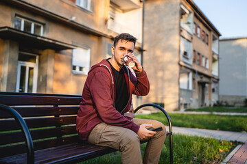 Obraz premium One young adult caucasian man sitting outdoor on the bench in autumn day holding mobile phone and a cup of coffee looking to the side alone waiting making a phone call or sms in front of building